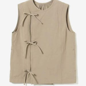 May include: A sleeveless, tan-colored top with a round neckline and three bow closures down the front. The top has vertical seams and a slightly gathered detail at the neckline. The fabric appears to be a lightweight material.
