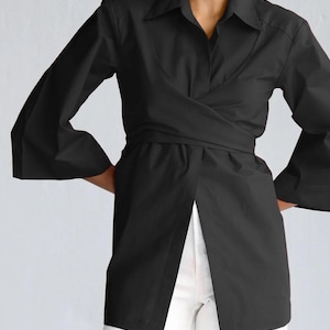Puede incluir: Una camisa negra envolvente con cuello y mangas largas acampanadas. La camisa presenta un diseño envolvente con cinturón en la cintura y abertura frontal. La modelo lleva pantalones blancos.