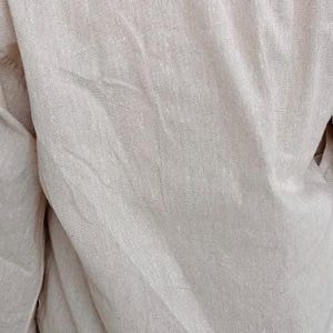 Puede incluir: Una camisa de manga larga en color beige claro con una textura tejida visible. La prenda presenta una costura horizontal en la parte superior de la espalda y muestra pliegues y arrugas naturales en toda la tela, resaltando el tejido.
