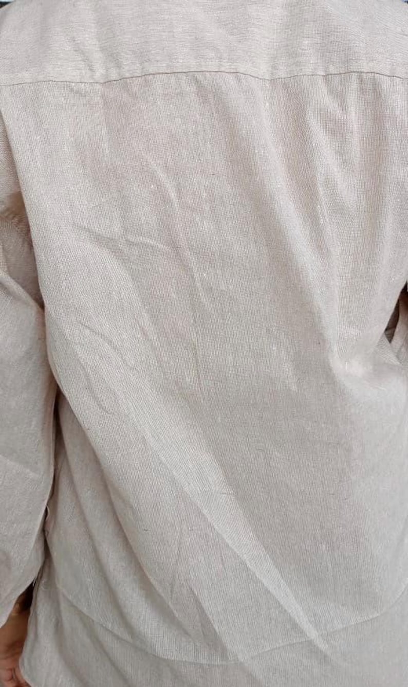 Puede incluir: Una camisa de manga larga en color beige claro con una textura tejida visible. La prenda presenta una costura horizontal en la parte superior de la espalda y muestra pliegues y arrugas naturales en toda la tela, resaltando el tejido.
