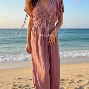 May include: A dusty rose maxi dress featuring a deep V-neckline, short flutter sleeves, and a braided waist detail. The garment has a textured fabric finish and includes the text GulaabCo and www.gulaabco.com printed on the lower portion.