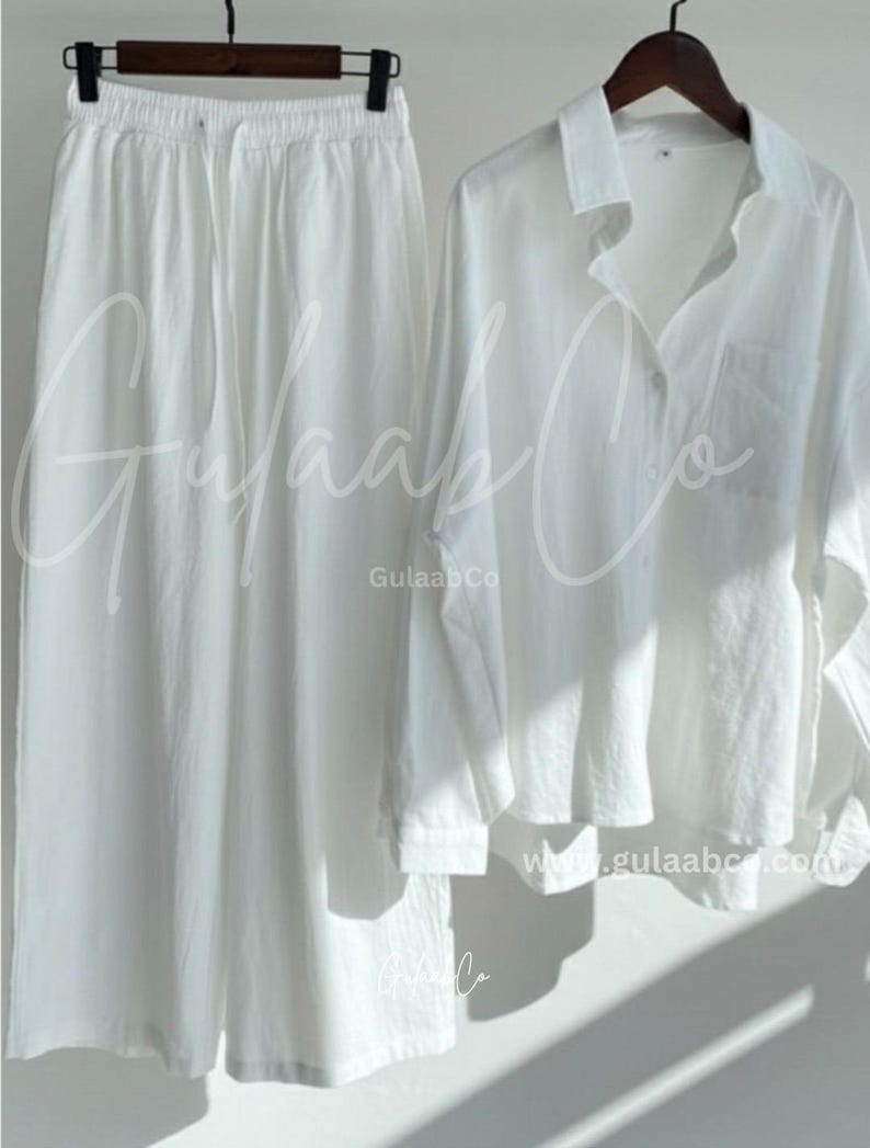 Puede incluir: Un conjunto de dos piezas blanco que consta de pantalones de pierna ancha con cintura el&aacute;stica con cord&oacute;n y una camisa de manga larga con botones y bolsillo en el pecho. El texto GulaabCo y www.gulaabco.com es visible en las prendas blancas.