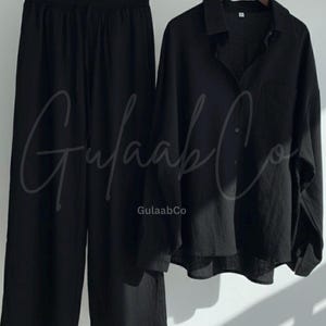 Puede incluir: Un conjunto negro de dos piezas que consta de una camisa de manga larga con botones, cuello y bolsillo en el pecho, junto con pantalones anchos con cintura el&aacute;stica y cord&oacute;n. El texto GulaabCo es visible en las prendas.