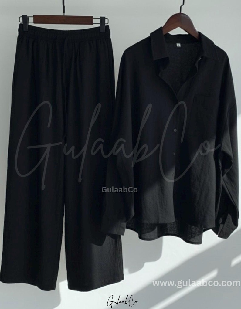 Puede incluir: Un conjunto negro de dos piezas que consta de una camisa de manga larga con botones, cuello y bolsillo en el pecho, junto con pantalones anchos con cintura el&aacute;stica y cord&oacute;n. El texto GulaabCo es visible en las prendas.