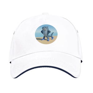 Pode incluir: Boné de beisebol branco com aba azul marinho e um gráfico de um tubarão de desenho animado. O tubarão é cinza e usa tênis azuis, em pé em uma praia com o oceano ao fundo. Um acessório casual.