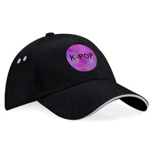 May include: Black baseball cap with a white trim on the brim. The cap features a circular design with the text "K-POP" in white letters on a pink and purple tie-dye background. The cap has a curved brim and a button on top.
