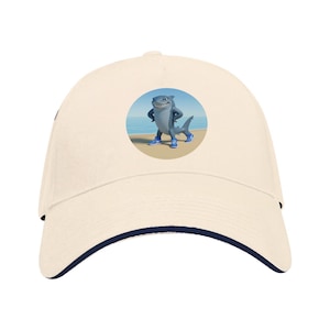 Pode incluir: Boné de beisebol creme com aba azul marinho. O boné apresenta um gráfico circular de um tubarão de desenho animado usando tênis azuis, em pé em uma praia com o oceano ao fundo. O tubarão tem uma expressão confiante.
