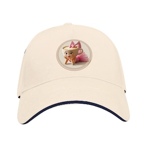 Pode incluir: Boné de beisebol creme com aba azul marinho. O boné apresenta um gráfico circular de um personagem de desenho animado em uma xícara de chá, usando um tutu e laço rosa. O personagem está apoiando o queixo nas mãos.