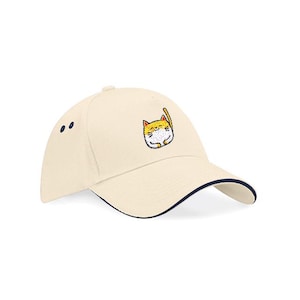 Op de afbeelding: Crèmekleurige baseballcap met een marineblauwe rand en een cartoonkat geborduurd op de voorkant. De kat is geel, wit en zwart. Twee marineblauwe stippen staan aan de zijkant van de pet.
