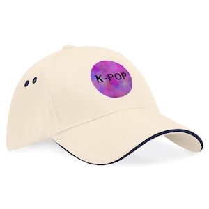 May include: Cream-colored baseball cap with a navy blue brim and accents. A circular patch on the front displays the text "K-POP" in white over a colorful, abstract design. The cap has a curved visor and a structured crown.