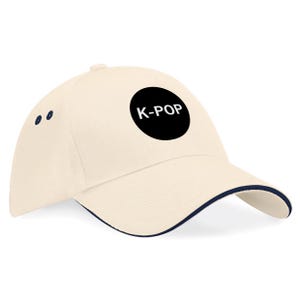 May include: Cream-colored baseball cap with a navy blue brim and accents. A black circle on the front displays the white text "K-POP". The cap has a curved visor and a button on top.