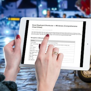 May include: A white tablet showing an Excel keyboard shortcuts cheat sheet for Windows. The screen displays text and a table of shortcuts. The tablet is held by a person with red nail varnish and a gold ring. The background is a blurred city street.