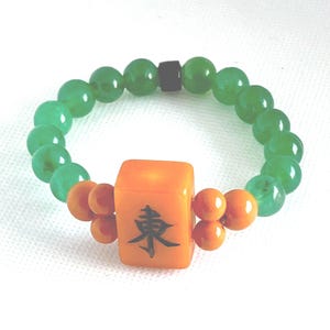 May include: A green beaded bracelet with an orange square centerpiece. The square features a black symbol. Small orange beads flank the centerpiece. The bracelet is a stretch style.