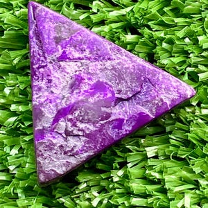 May include: A triangular, polished purple gemstone with a textured surface. The stone has a mix of light and dark purple hues and is set against a backdrop of green artificial grass.