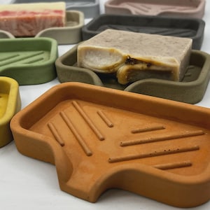 May include: A collection of colorful soap dishes in various shades, including yellow, green, and brown. Each dish holds a bar of soap. The soap dishes have a rectangular shape with a sloped design and are made of a solid material.