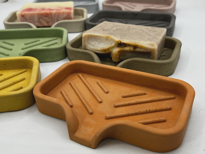 May include: A collection of colorful soap dishes in various shades, including yellow, green, and brown. Each dish holds a bar of soap. The soap dishes have a rectangular shape with a sloped design and are made of a solid material.
