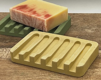 May include: A rectangular bar of soap with a marbled cream and red pattern resting on a matte olive green soap dish, positioned behind a second empty soap dish in a muted mustard yellow color, both featuring a slatted design for drainage.