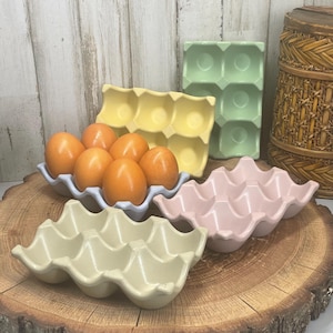 May include: A collection of colorful ceramic egg holders in pastel shades of yellow, green, pink, and cream. One holder contains six brown eggs. The egg holders are arranged on a wooden surface, suggesting a rustic or farmhouse aesthetic.