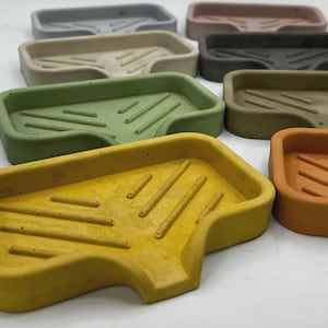 May include: A collection of rectangular soap dishes in various colors, including gray, beige, green, yellow, and orange. Each dish has a sloped design with grooves for drainage. The soap dishes are made of a solid material.