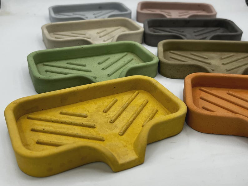 May include: A collection of rectangular soap dishes in various colors, including gray, beige, green, yellow, and orange. Each dish has a sloped design with grooves for drainage. The soap dishes are made of a solid material.