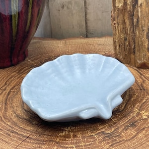 May include: A light blue ceramic seashell-shaped dish. The dish has a scalloped edge and a textured surface. The dish is sitting on a wooden surface.