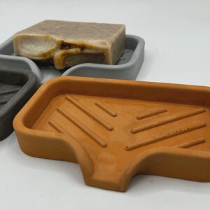 May include: Three concrete soap dishes in gray and orange. The orange dish is in the foreground, with a rectangular shape and diagonal grooves. A gray dish and a gray soap dish with a bar of soap are in the background.