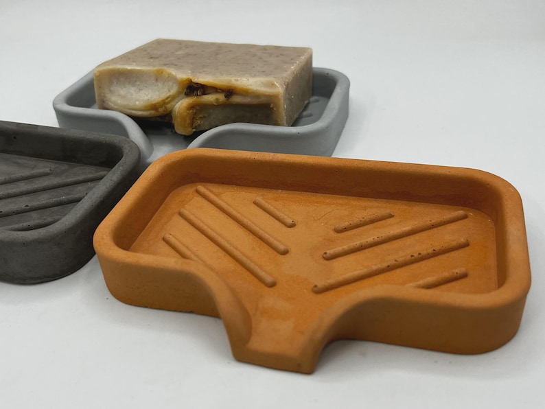 May include: Three concrete soap dishes in gray and orange. The orange dish is in the foreground, with a rectangular shape and diagonal grooves. A gray dish and a gray soap dish with a bar of soap are in the background.