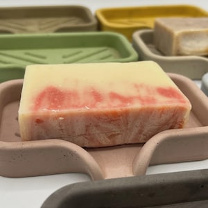 May include: A close-up of several soap dishes in various colors, including green, yellow, and gray. A bar of soap with red and white marbling sits in a pink dish. The soap dishes are made of a solid material.