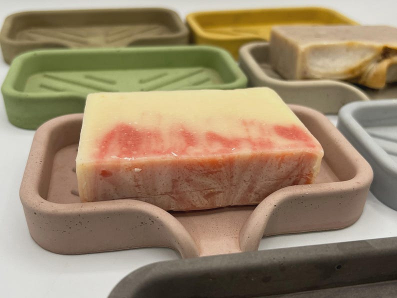 May include: A close-up of several soap dishes in various colors, including green, yellow, and gray. A bar of soap with red and white marbling sits in a pink dish. The soap dishes are made of a solid material.