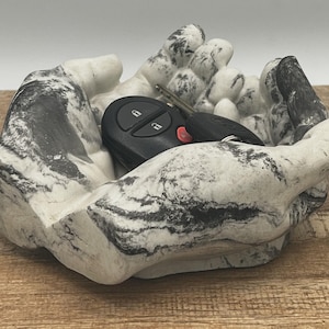May include: A marble-like decorative bowl in the shape of cupped hands, with black and gray veining. A black car key with a red button rests inside the bowl. The bowl is on a wooden surface.