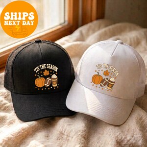May include: Two baseball caps, one black and one white, each featuring a graphic with a pumpkin, a football, and a coffee cup. The text 'TIS THE SEASON' is printed in an arc above the illustrations, surrounded by small stars and a leaf.