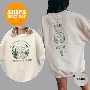 May include: A sand-colored crewneck sweatshirt featuring a dark green graphic print. The front displays a mountain landscape inside a laurel wreath with the text "PROTECT OUR NATIONAL PARK" and "PARKS OVER PROFIT". The back shows a vertical nature design.