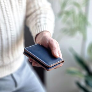 Può includere: Un portafoglio in pelle blu navy con cuciture bianche e un bordo marrone chiaro. Il portafoglio è tenuto in mano da una persona, con uno sfondo sfocato di un maglione bianco e verde.