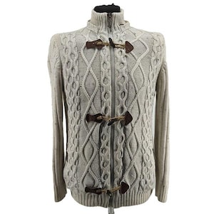 May include: A light beige cable-knit sweater with a high collar and a full zipper. The sweater features three toggle closures made of brown leather and rope. The sleeves are long, and the overall design is a classic, textured knit.