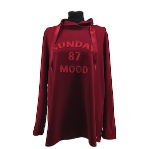 Könnte beinhalten: Burgunderfarbener Hoodie mit langen Ärmeln und dem gestickten Text "SUNDAY 87 MOOD" auf der Vorderseite. Der Hoodie hat eine Kapuze mit Kordelzug und wird auf einer Schaufensterpuppe präsentiert. Der Stoff wirkt weich und bequem.