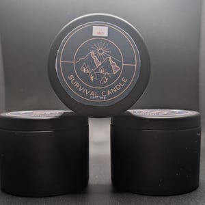 Könnte beinhalten: Drei schwarze Überlebenskerzen mit einem kreisförmigen Etikett mit einem Berg- und Sonnendesign. Das Etikett lautet "SURVIVAL CANDLE" und "95 g". Die Kerzen befinden sich in schwarzen Dosen.