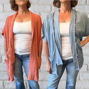 May include: Two open-front cardigans, one coral and one light blue, with short sleeves and large front pockets. The coral cardigan is made of a textured fabric. The light blue cardigan has sequin trim along the edges. Both are worn over a white tank top and blue jeans.