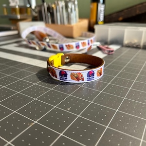 May include: A brown leather bracelet with a white ribbon featuring Taco Bell logos. The bracelet has a yellow clasp. Another similar bracelet is visible in the background, along with crafting tools and supplies.