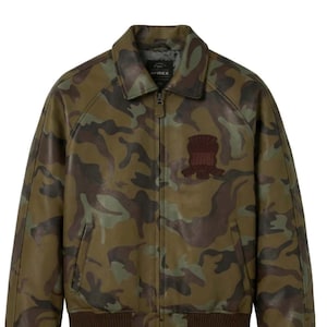 Puede incluir: Chaqueta de cuero con estampado de camuflaje en tonos verdes, marrones y beige. La chaqueta tiene cuello, cierre de cremallera y puños y dobladillo de canalé marrón. Un parche marrón está en el pecho. La marca "AVIREX" es visible.