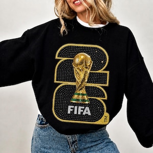 Op de afbeelding: Zwart sweatshirt met een gouden en strass ontwerp met het nummer 2022, een gouden trofee en het woord FIFA in het wit. De sweatshirt heeft lange mouwen en een ronde hals.