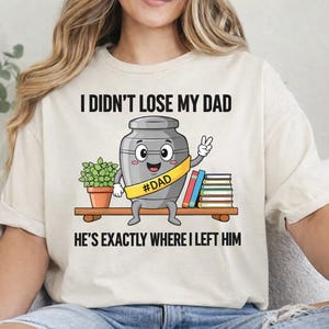 Puede incluir: Camiseta color crema con el texto "I Didn't Lose My Dad, He's Exactly Where I Left Him". El gráfico presenta una urna de dibujos animados con una banda #DAD, una planta y libros en un estante. La urna hace el signo de la paz.