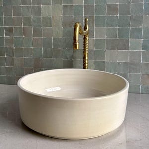 Könnte beinhalten: Ein rundes, cremefarbenes Keramik-Aufsatzwaschbecken mit glatter, glänzender Oberfläche. Hinter dem Becken befindet sich eine hohe, vertikale Messingarmatur mit einem strukturierten, gerippten zylindrischen Schaft und einem gebogenen Auslauf.