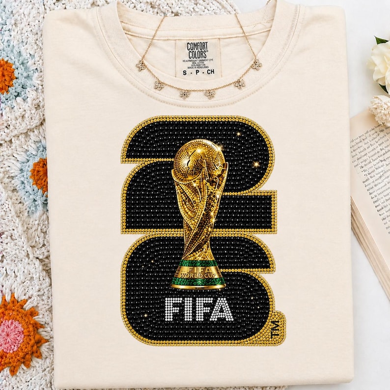 Op de afbeelding: Cr&egrave;mekleurig T-shirt met een grote afbeelding van de FIFA World Cup-trofee. De afbeelding is omlijnd met gouden en zwarte strass-steentjes, met het woord "FIFA" in witte strass-steentjes. Een gouden ketting met kleine bloemhangers is op het shirt gedrapeerd.