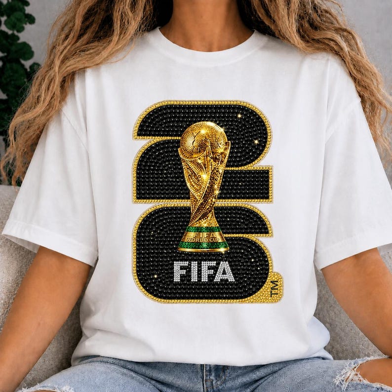 Op de afbeelding: Wit T-shirt met een gouden FIFA World Cup trofee ontwerp. Het ontwerp bevat de tekst "FIFA" in zilveren letters, tegen een zwarte achtergrond met gouden accenten. Het shirt heeft een klassieke ronde hals.