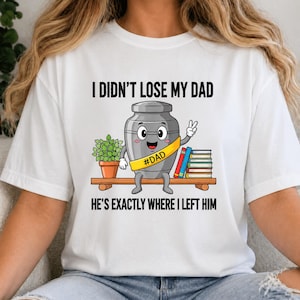 Puede incluir: Camiseta blanca con el texto "I Didn't Lose My Dad He's Exactly Where I Left Him." El gráfico muestra una urna de dibujos animados con una faja amarilla que dice "#DAD" sentada en un estante con una planta y libros.