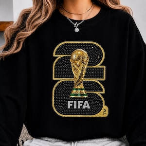 Op de afbeelding: Zwarte sweatshirt met een gouden FIFA World Cup trofee ontwerp. Het nummer "2022" is omlijnd met gouden en zwarte strass-steentjes, met het woord "FIFA" in het wit aan de onderkant. Het ontwerp is gecentreerd op de voorkant.