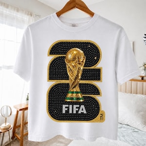 Op de afbeelding: Wit T-shirt met een gouden FIFA World Cup trofee ontwerp. Het ontwerp bevat het nummer 23 in zwart met gouden en witte accenten, en het woord "FIFA" in wit. Het shirt hangt aan een houten hanger.