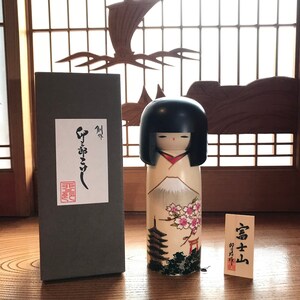 May include: A traditional Japanese kokeshi doll with a black hairpiece and a painted face. The doll is wearing a white kimono with a red sash and a mountain scene with cherry blossoms and a pagoda. The doll is in a box with Japanese writing on it.