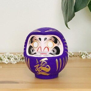 May include: A purple and gold Japanese lucky charm, a daruma doll, with a white face and gold accents. The doll is sitting on a wooden surface with a white background.
