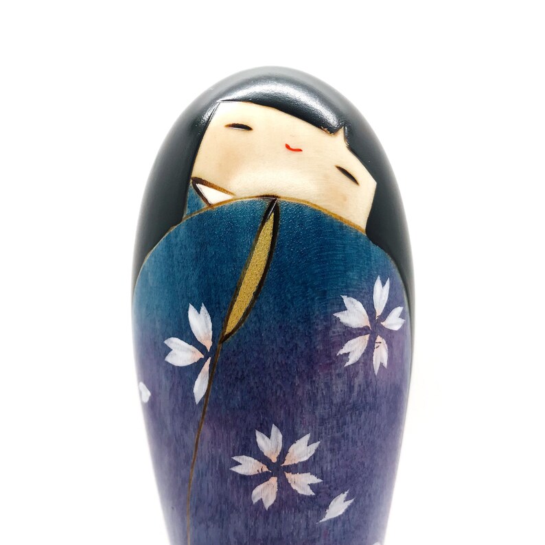 May include: A wooden doll with a black hair, a purple and blue kimono with white flower designs, and a smiling face.